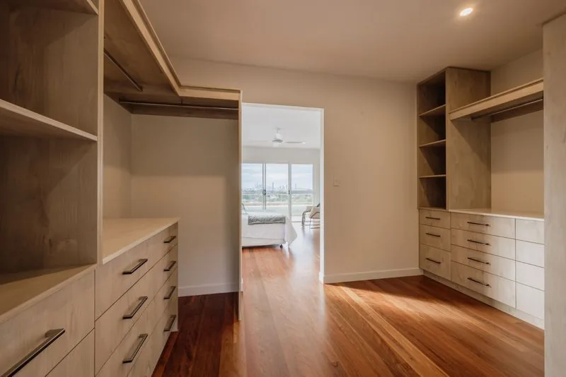 Walk-in robe in timber joinery, with banks of drawers and open shelving, looking through to the bedroom
