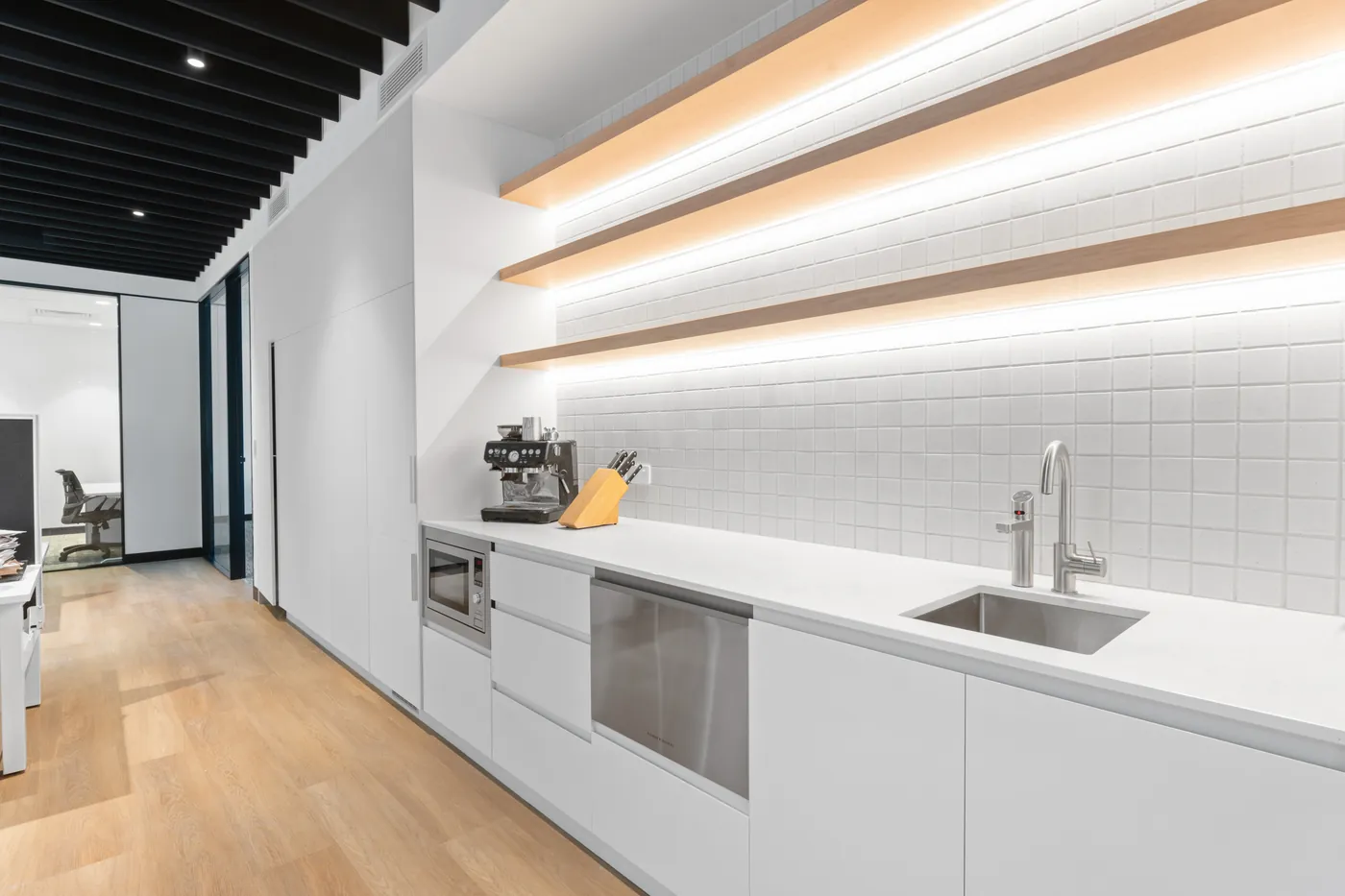Office kitchenette at the Sumner fit-out with white cabinetry, LED-lit timber shelves and a tiled splashback