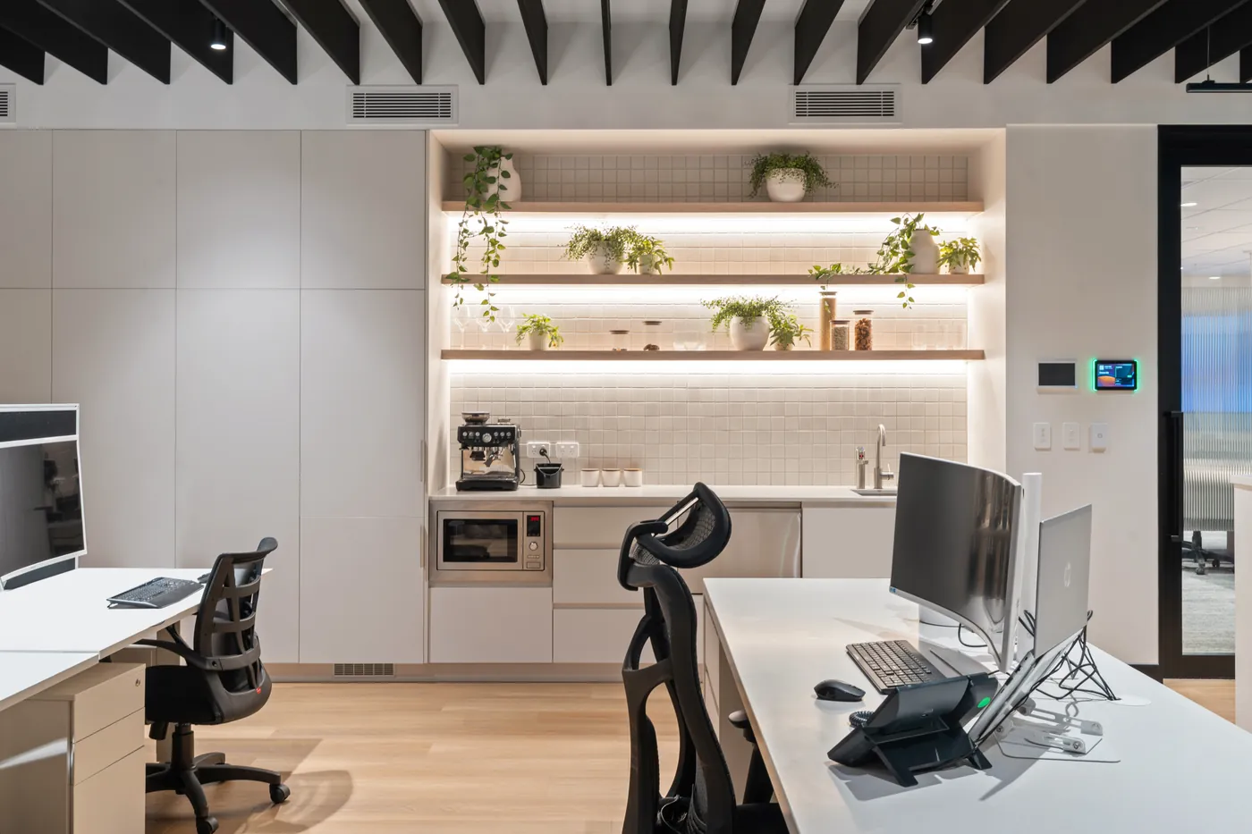 Office kitchenette with white cabinetry, LED-lit timber shelves and a tiled splashback beside the workstations