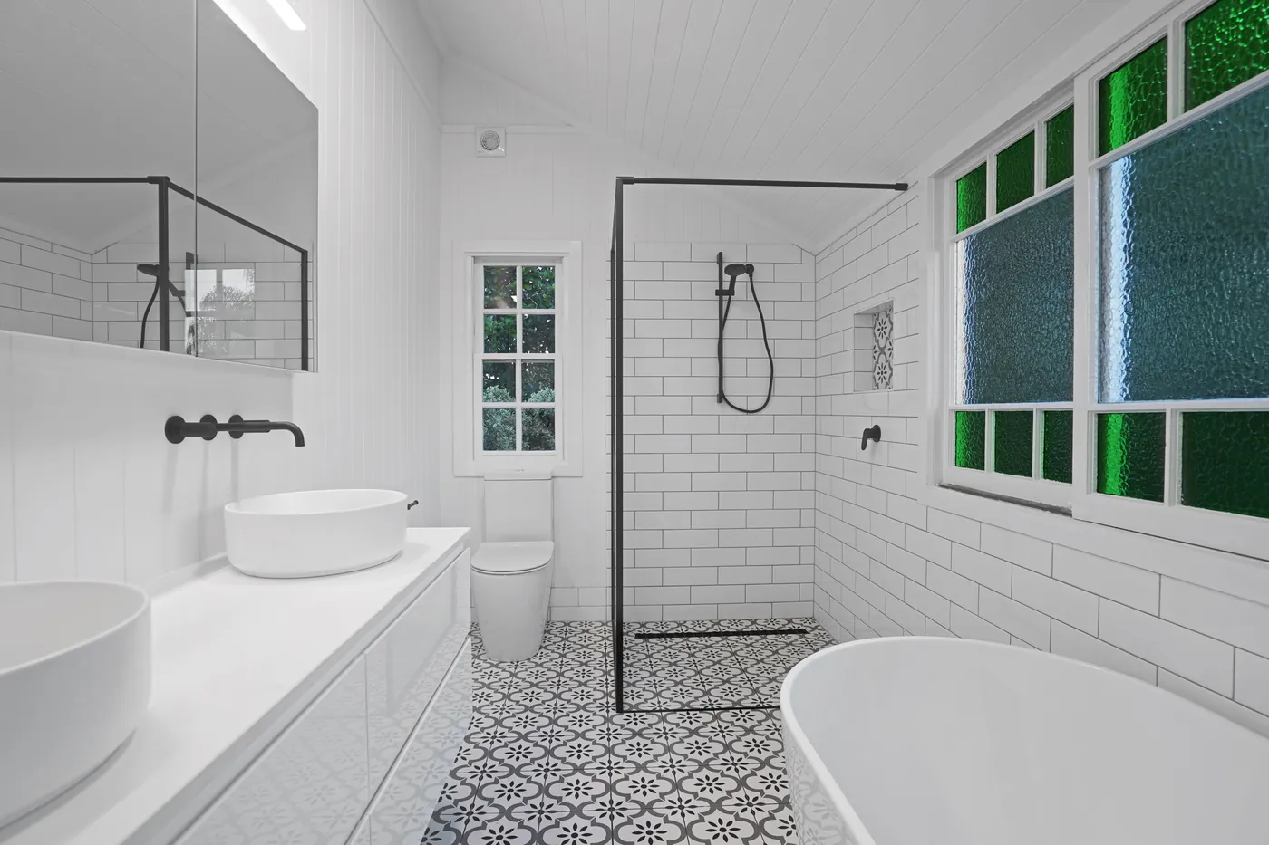 Renovated bathroom with a vessel basin, walk-in shower in black tapware, freestanding bath and patterned floor tiles beneath green and blue leadlight windows